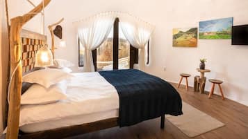 A bedroom with a wooden headboard, a bed with white and dark bedding, a bedside lamp, a large window with curtains, and two paintings on the wall.