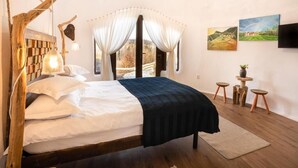 A bedroom with a wooden headboard, a bed with white and dark bedding, a bedside lamp, a large window with curtains, and two paintings on the wall.