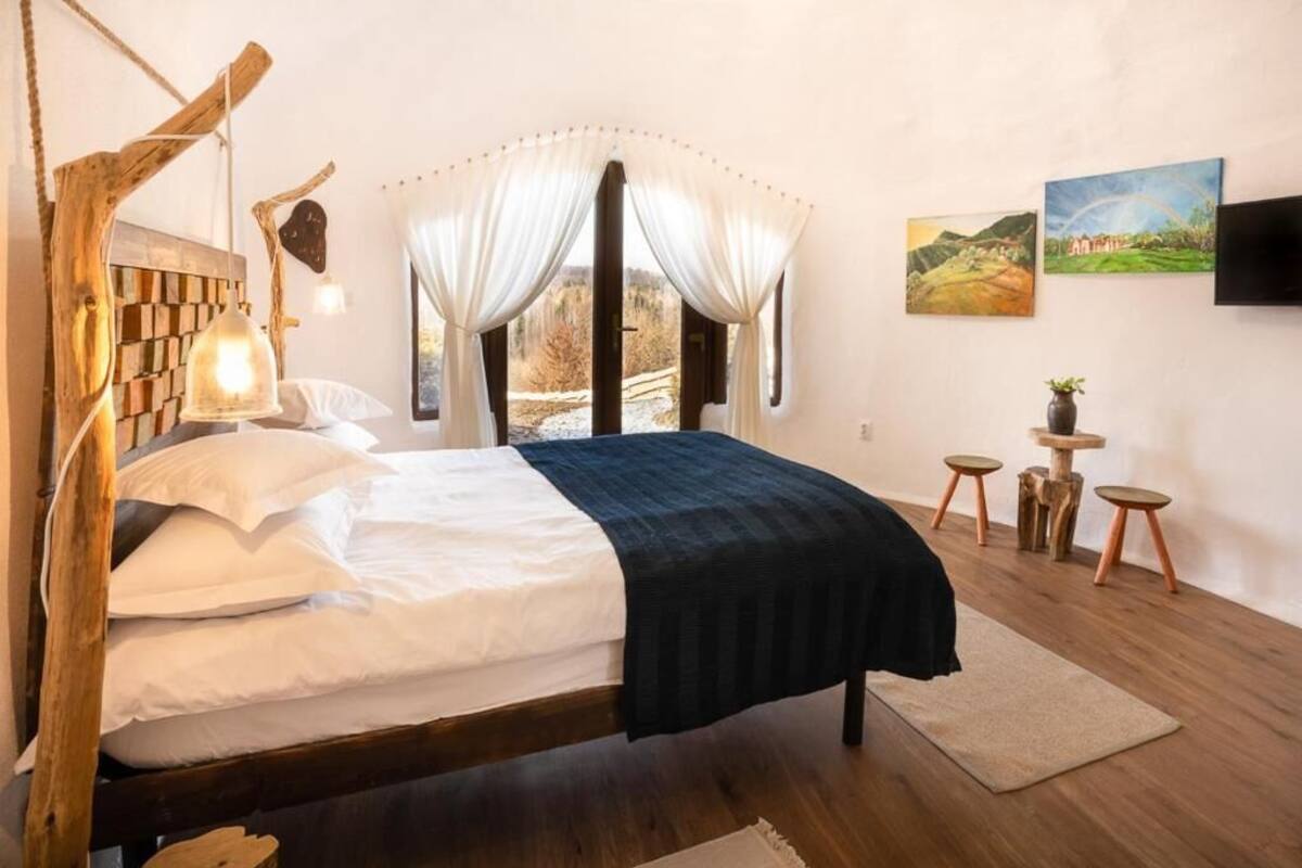 Un dormitorio con cabecera de madera, cama con ropa de cama blanca y oscura, una lámpara de noche, un ventanal con cortinas y dos cuadros en la pared.