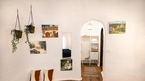 Un pasillo con suelo de madera, paredes blancas y varias obras de arte. Hay un banco blanco con cojines, una planta colgante y un estante blanco con objetos encima.