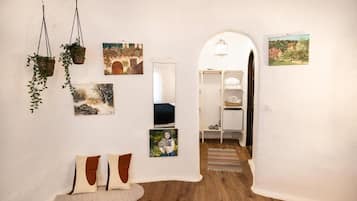 A hallway with a wooden floor, white walls, and various artworks. There is a white bench with cushions, a hanging plant, and a white shelving unit with items on it.