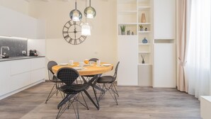 Uma zona de refeições moderna com uma mesa redonda de madeira, cadeiras de estrutura de fio preto e duas luzes pendentes. Um relógio de parede grande e uma estante branca com objetos decorativos também são visíveis.