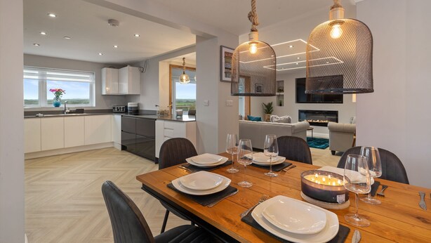 A modern dining area with a wooden table set for a meal, a chandelier, and a view of the kitchen and living area. - Golwg Y M r - Sea View Cottage With Hot Tub (Saundersfoot)