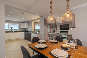 A modern dining area with a wooden table set for a meal, a chandelier, and a view of the kitchen and living area.