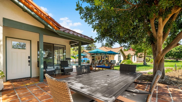 Une terrasse extérieure avec une table en bois et des chaises en osier, un espace salon abrité et une vue sur une maison en arrière-plan.