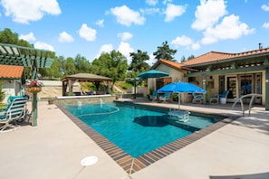 A swimming pool with lounge chairs and umbrellas, a covered patio area, and a building with a tiled roof.