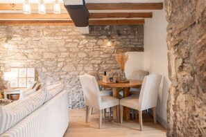 Un interior acogedor con una pared de piedra, vigas de madera, una mesa redonda de madera con sillas, un sofá con cojines y una lámpara colgante.