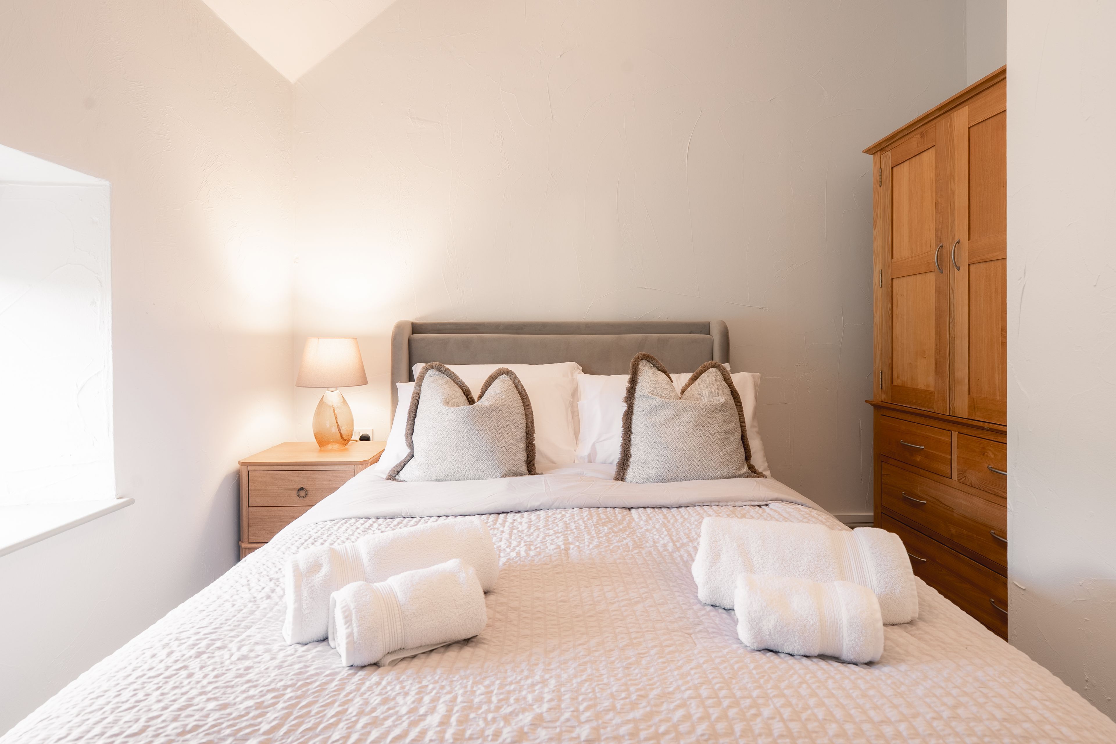 A neatly made bed with white linens and two towels, a bedside lamp, and a wooden wardrobe.