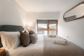 Ein Schlafzimmer mit einem Bett, Kissen, einem Nachttisch, einer Lampe, einem Fenster mit Vorhängen und einem Spiegel an der Wand.