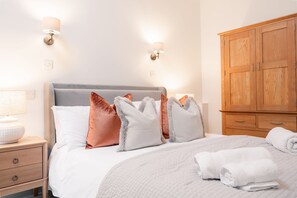 A neatly made bed with a grey headboard, multiple pillows, and a white blanket. There is a wooden wardrobe to the right and a bedside table with a lamp on the left. - Walker's Rest Cottage - perfect for two in the Peaks (Buxton)