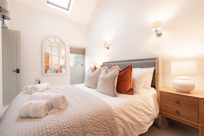 Un dormitorio con cama, almohadas, una mesita de noche, una lámpara, una ventana y una puerta.