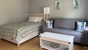 A bedroom with a bed, a grey sofa, a white coffee table, and a ceiling fan. - Studio steps to a private beach!! Ocean views from backyard!! 2 Paddle Boards!!  (Dennis)