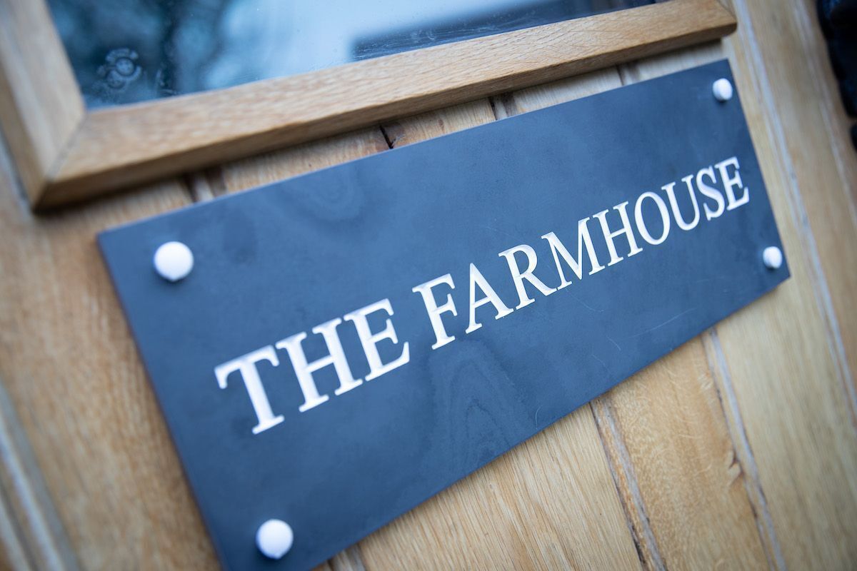 Een donkerblauwen bord met de woorden 'THE FARMHOUSE' in witte, hoofdletters, bevestigd aan een houten oppervlak met zichtbare schroeven.