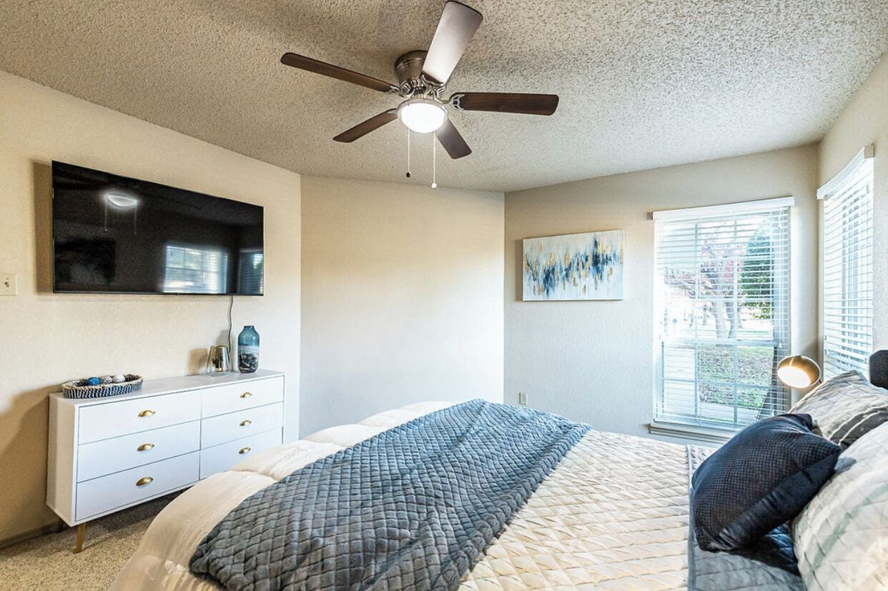 Une chambre à coucher comprenant un lit, un ventilateur de plafond, une télévision, une commode et une fenêtre avec des stores.
