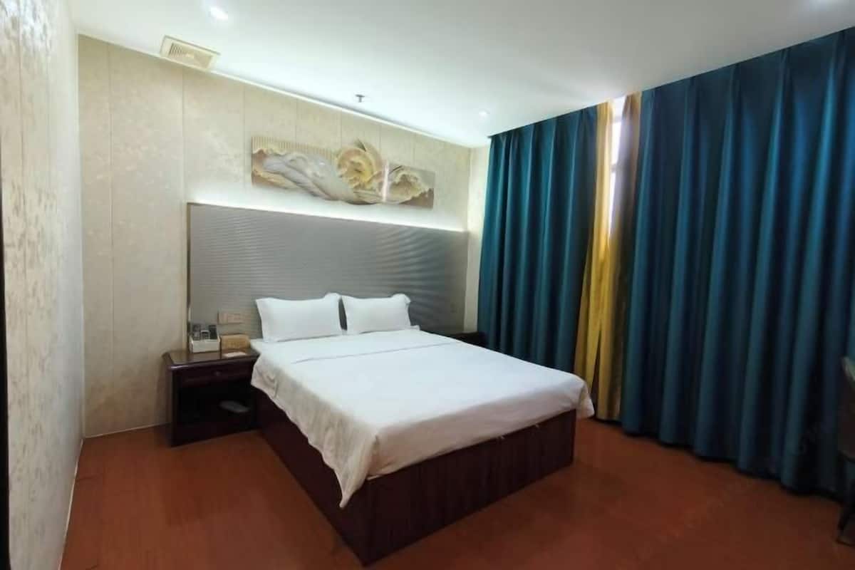 A hotel room with a large bed, a nightstand, a wall-mounted artwork, and a window with curtains.