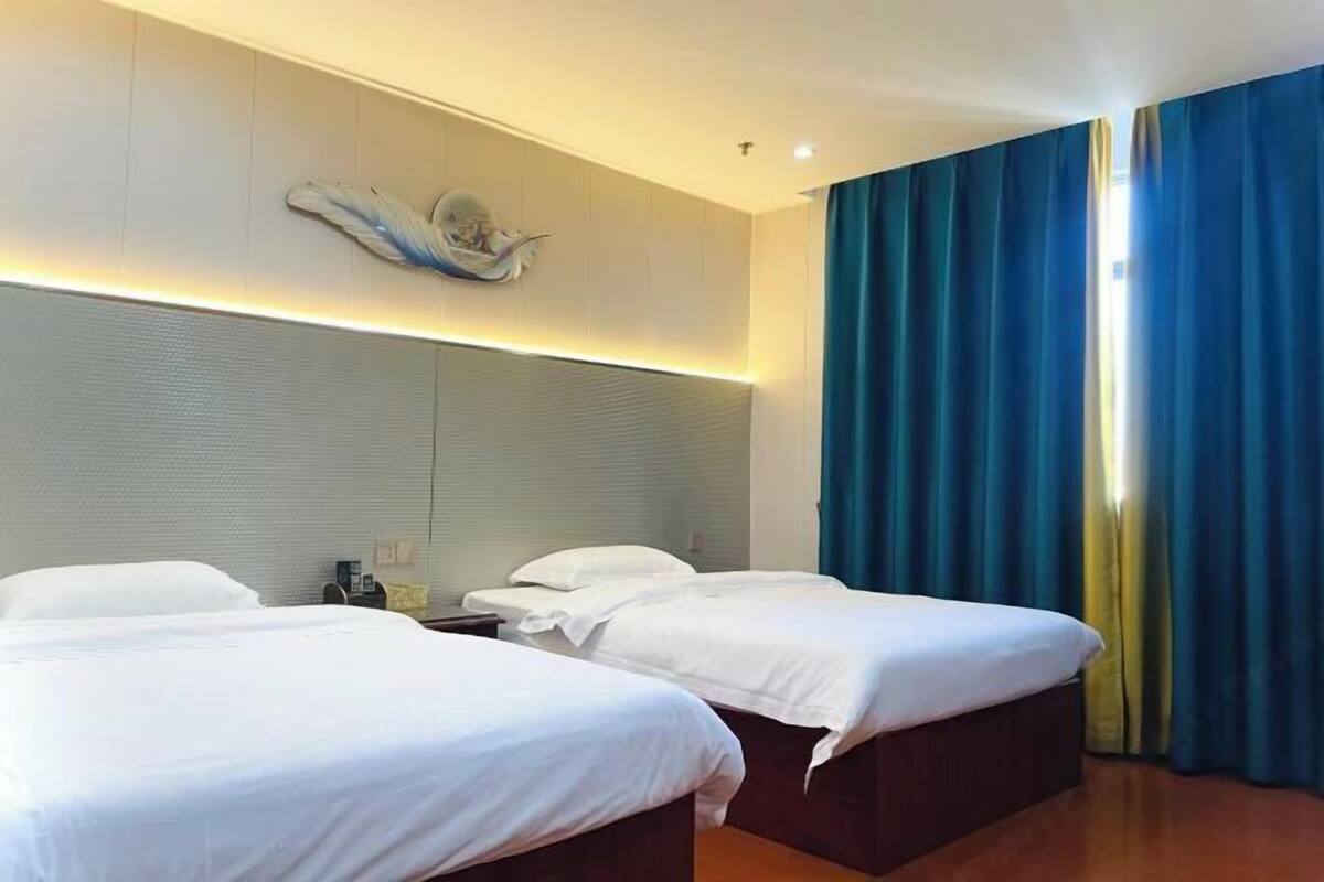 A hotel room with two beds, a large wall-mounted artwork, and a view of the outdoors through a window with blue curtains.