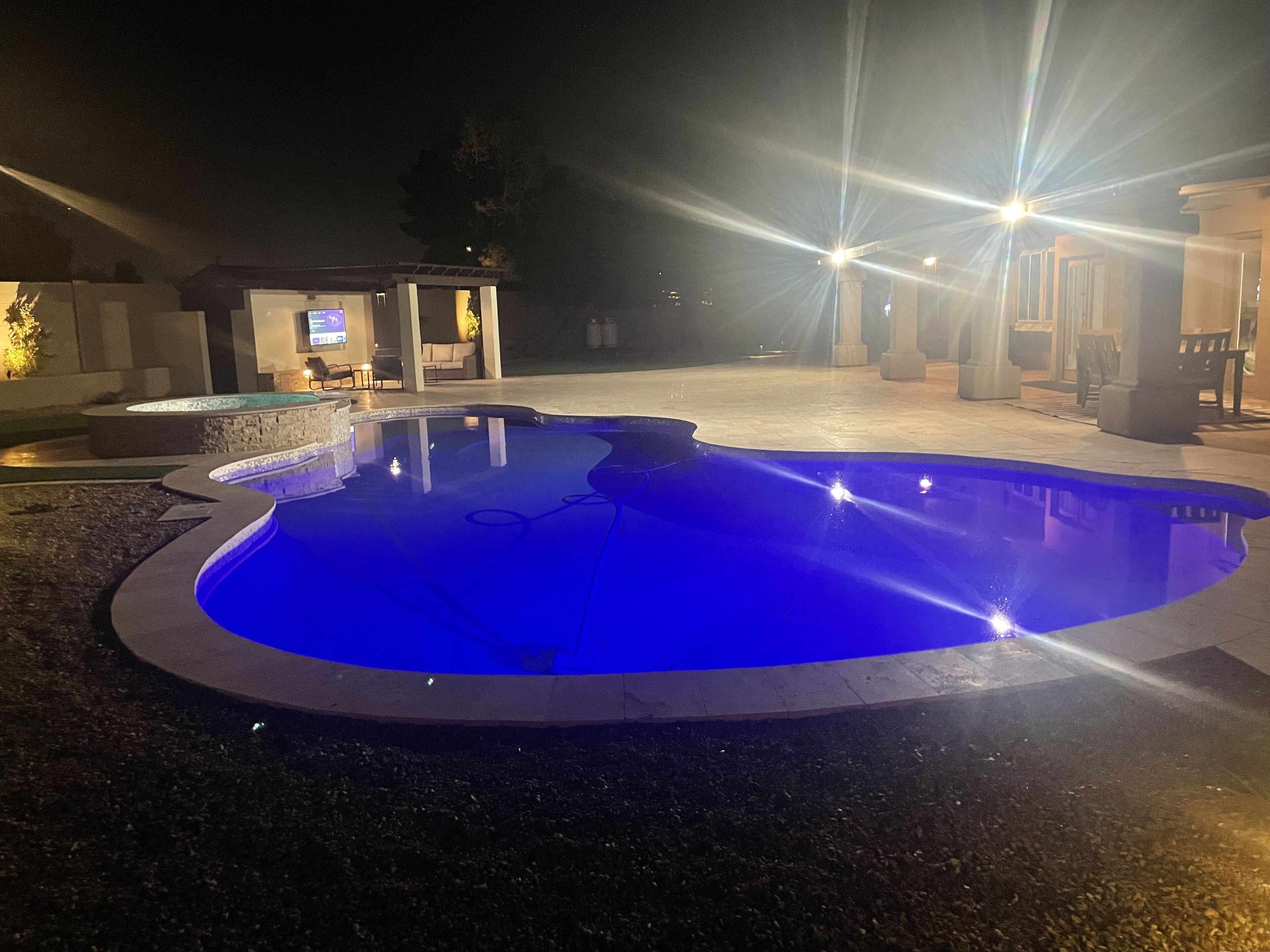 An outdoor swimming pool with a blue tint, surrounded by a stone deck and a small seating area with a TV.
