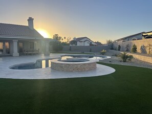 Un patio trasero con jardines, un elemento decorativo circular de piedra, una piscina y una casa al fondo.