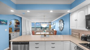 A kitchen with white cabinets, a dishwasher, and a microwave. There is a dining area with a table and chairs, and a view of the ocean through large windows.