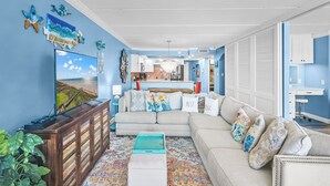 A living room with a sofa, a coffee table, a TV, and a kitchen in the background.