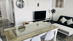 A modern living room with a glass-top dining table, a flat-screen TV, a black sofa with patterned cushions, and a standing fan. - Private Spacious villa  Ocean view and Rooftop Bar. Tourism Authority licence. (Flic en Flac)