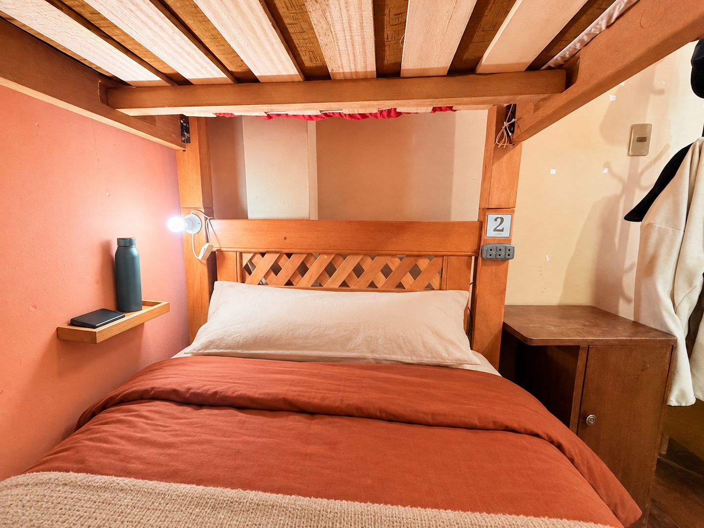 A single bed with a wooden headboard, a bedside table, a wall-mounted shelf, and a number indicating room 2.
