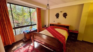 A bedroom with a large window, a bed with a red blanket, a wooden nightstand, and a small table with a vase and flowers. - Casa de huespedes La Casa Bonita (La Paz)
