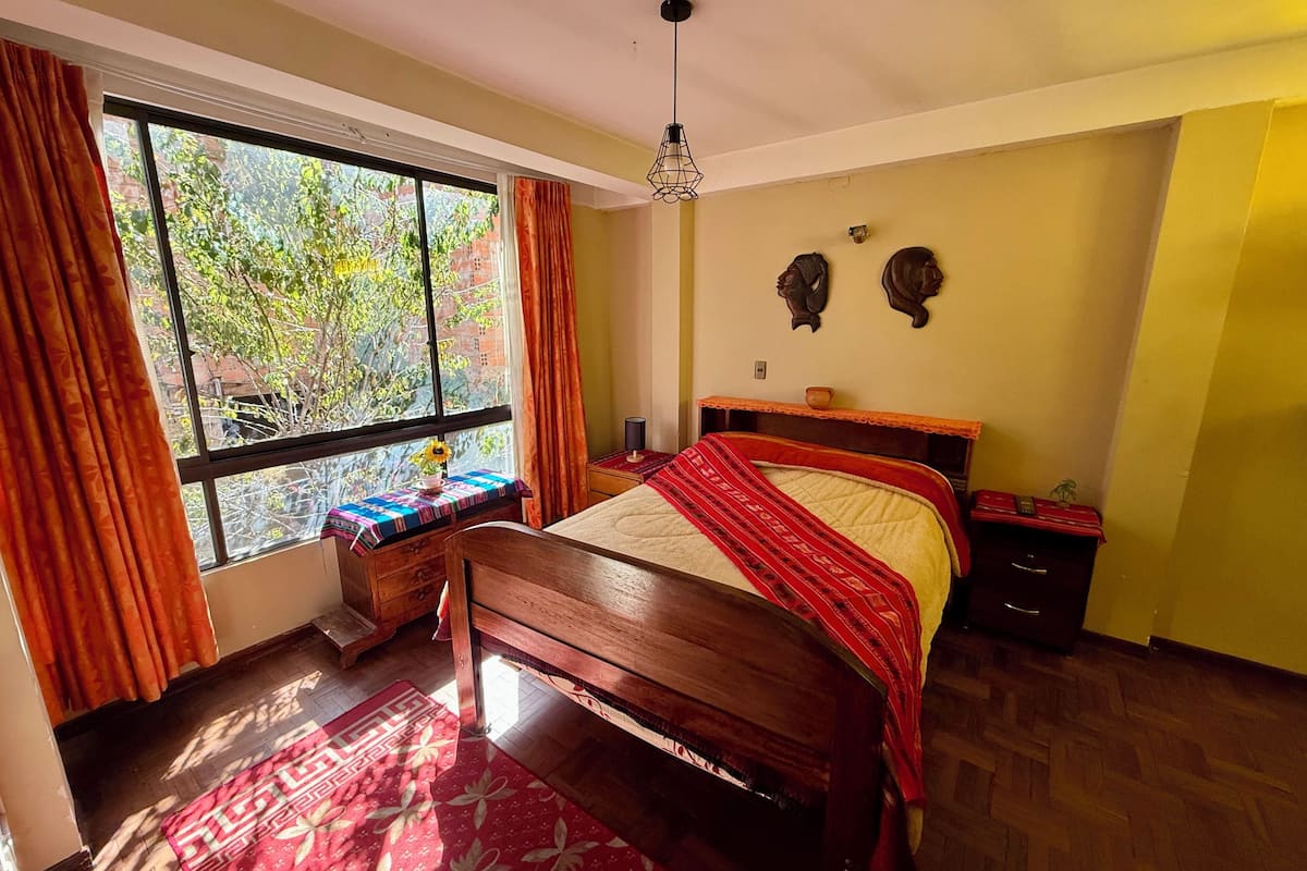 Ein Schlafzimmer mit großem Fenster, einem Bett mit roter Decke, einem Nachttisch aus Holz und einem kleinen Tisch mit Vase und Blumen.