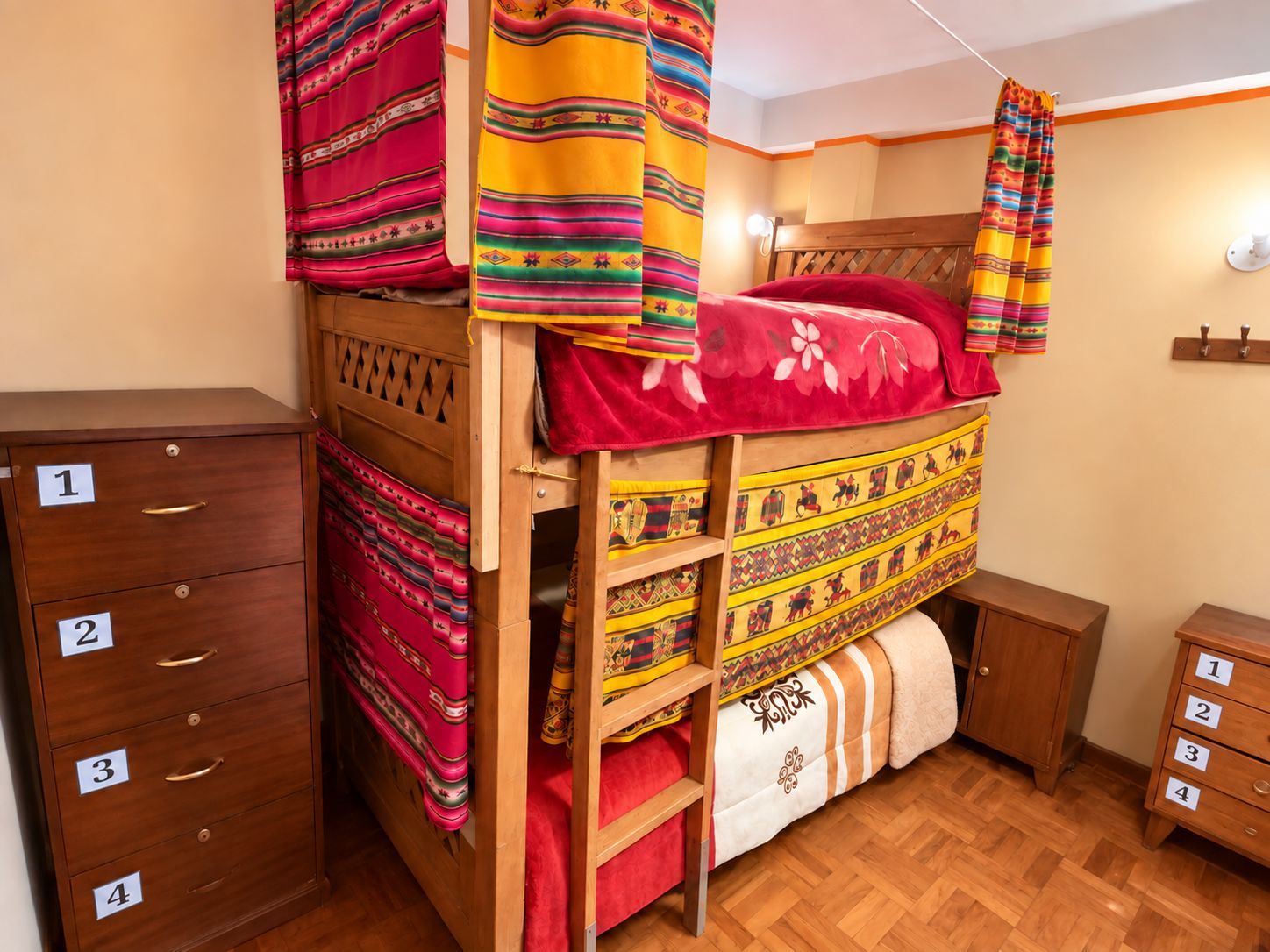 Een stapelbed met kleurrijke dekens, een houten kast met genummerde lades en een klein nachtkastje.