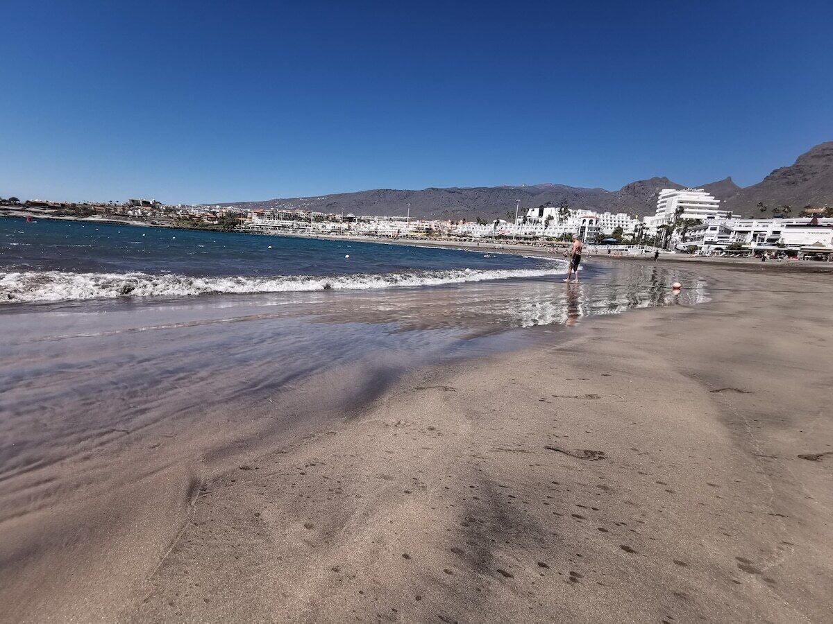Una playa con cielo despejado, una persona caminando y edificios al fondo.