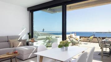 Un salón moderno con un sofá blanco, una mesa de comedor y sillas. Se aprecia una ventana grande con vistas al mar.
