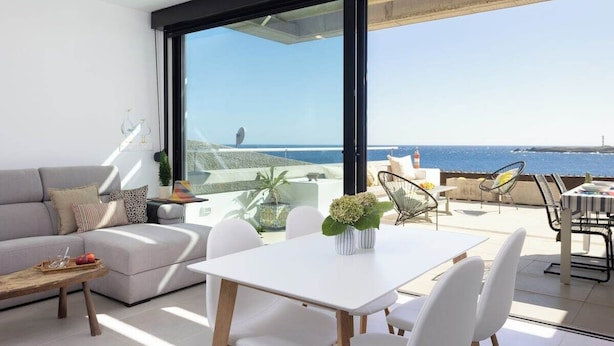 Un salón moderno con un sofá blanco, una mesa de comedor y sillas. Se aprecia una ventana grande con vistas al mar.