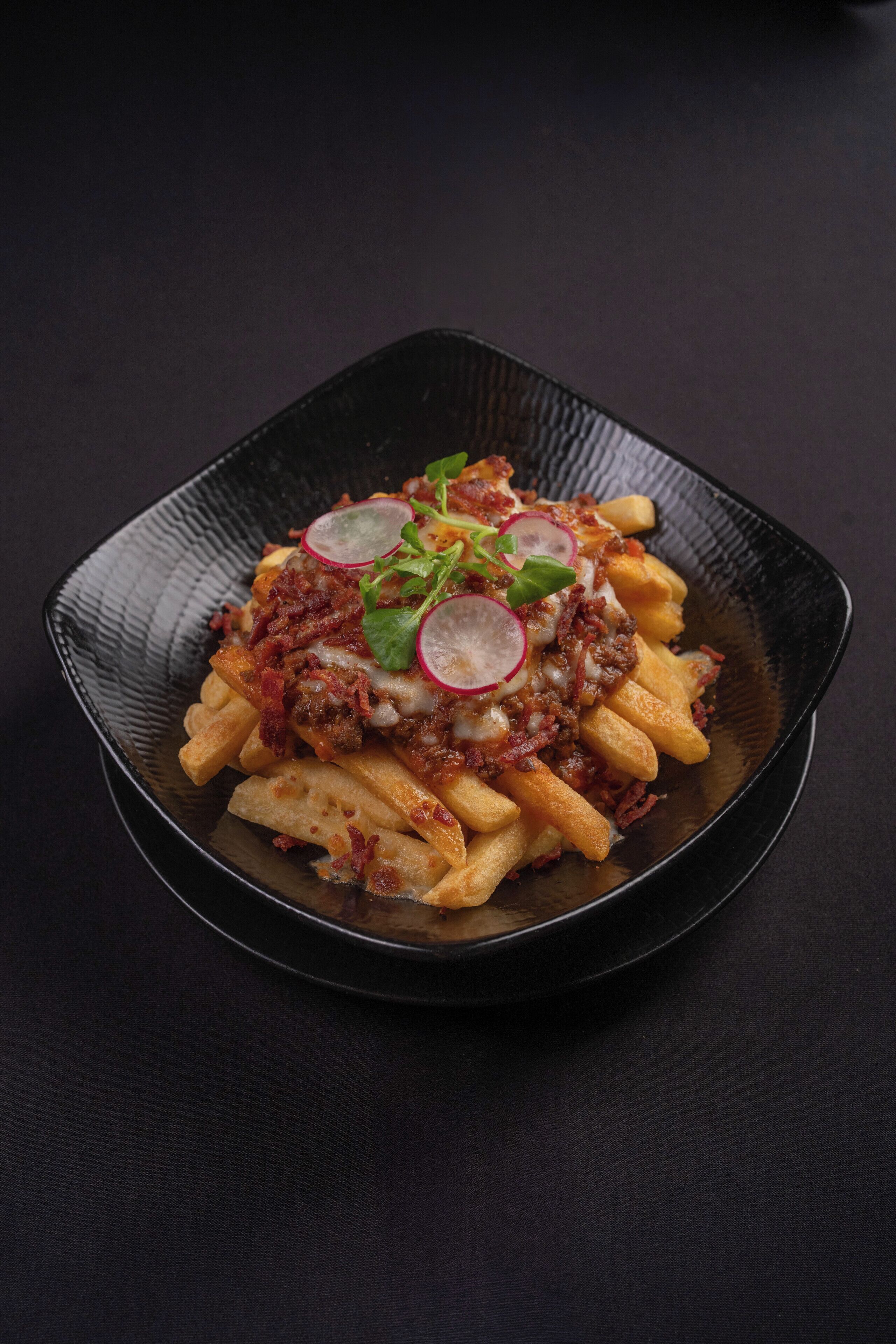 A square black plate with fries topped with meat, cheese, and radishes.