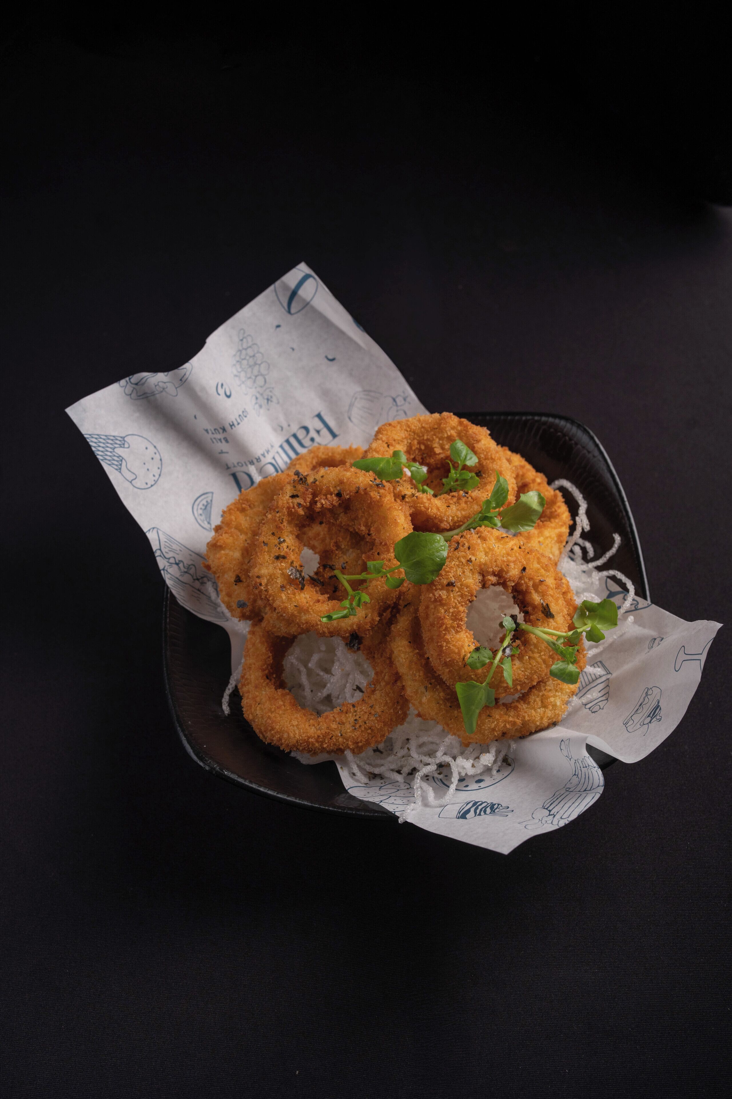 A basket of fried onion rings on a black tray with a white paper liner.
