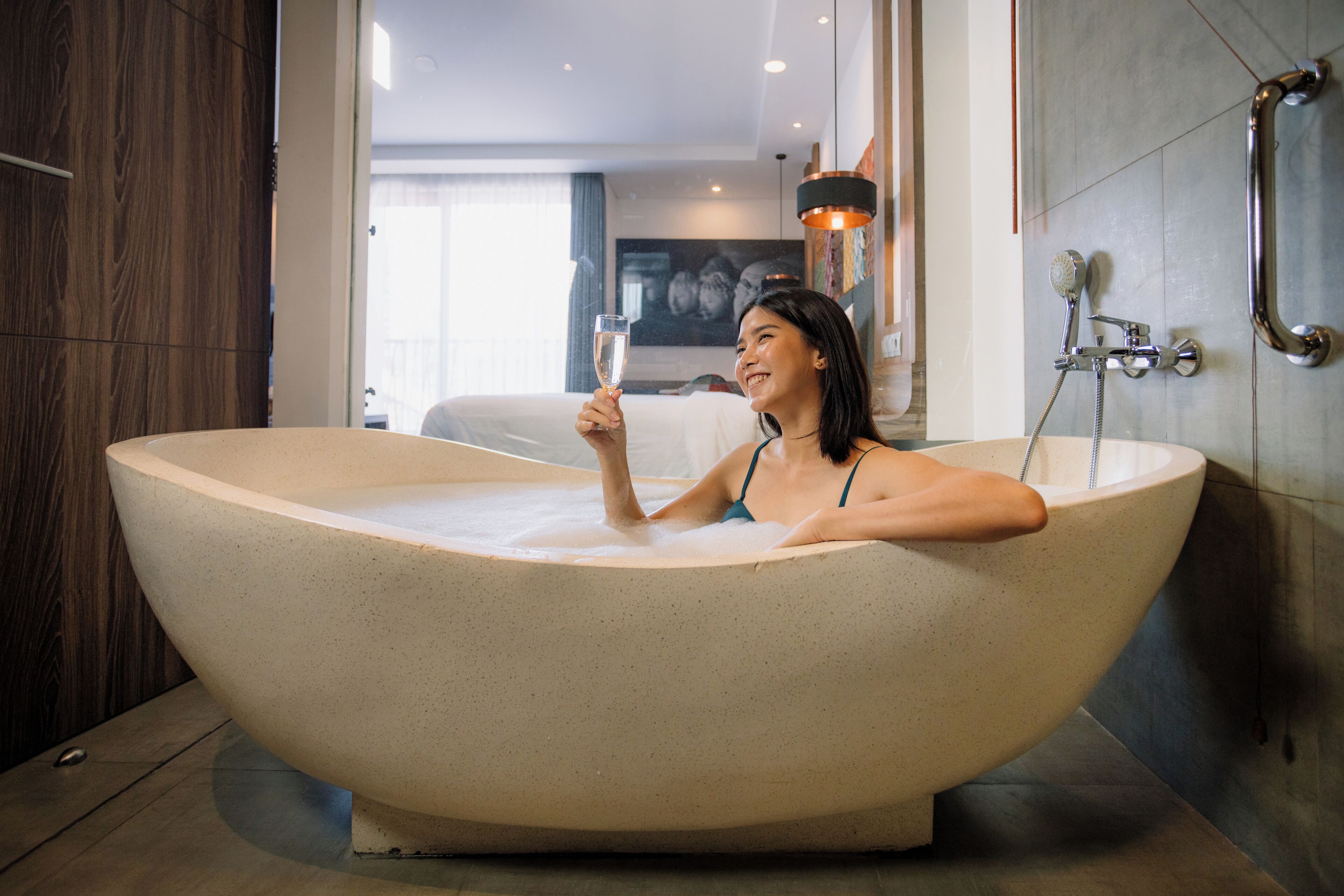 A woman in a large bathtub with a champagne glass, a showerhead, and a towel rack in a modern bathroom.