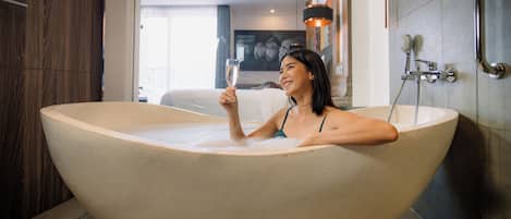 Una mujer en una bañera grande con una copa de champán, un cabezal de ducha y un perchero en un baño moderno.
