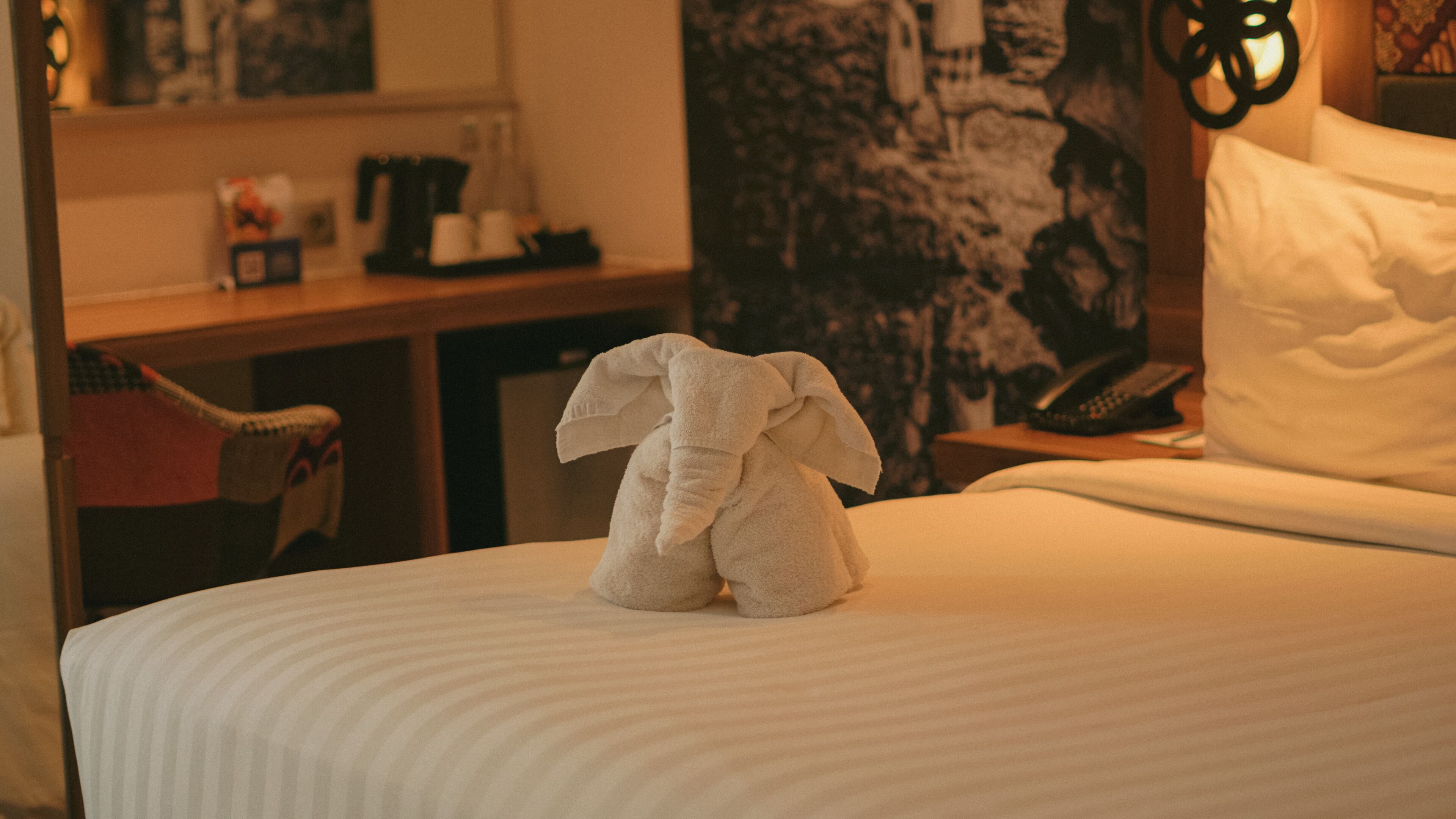 A neatly made bed with a towel shaped like an elephant, a telephone, and a wall with a patterned tapestry.