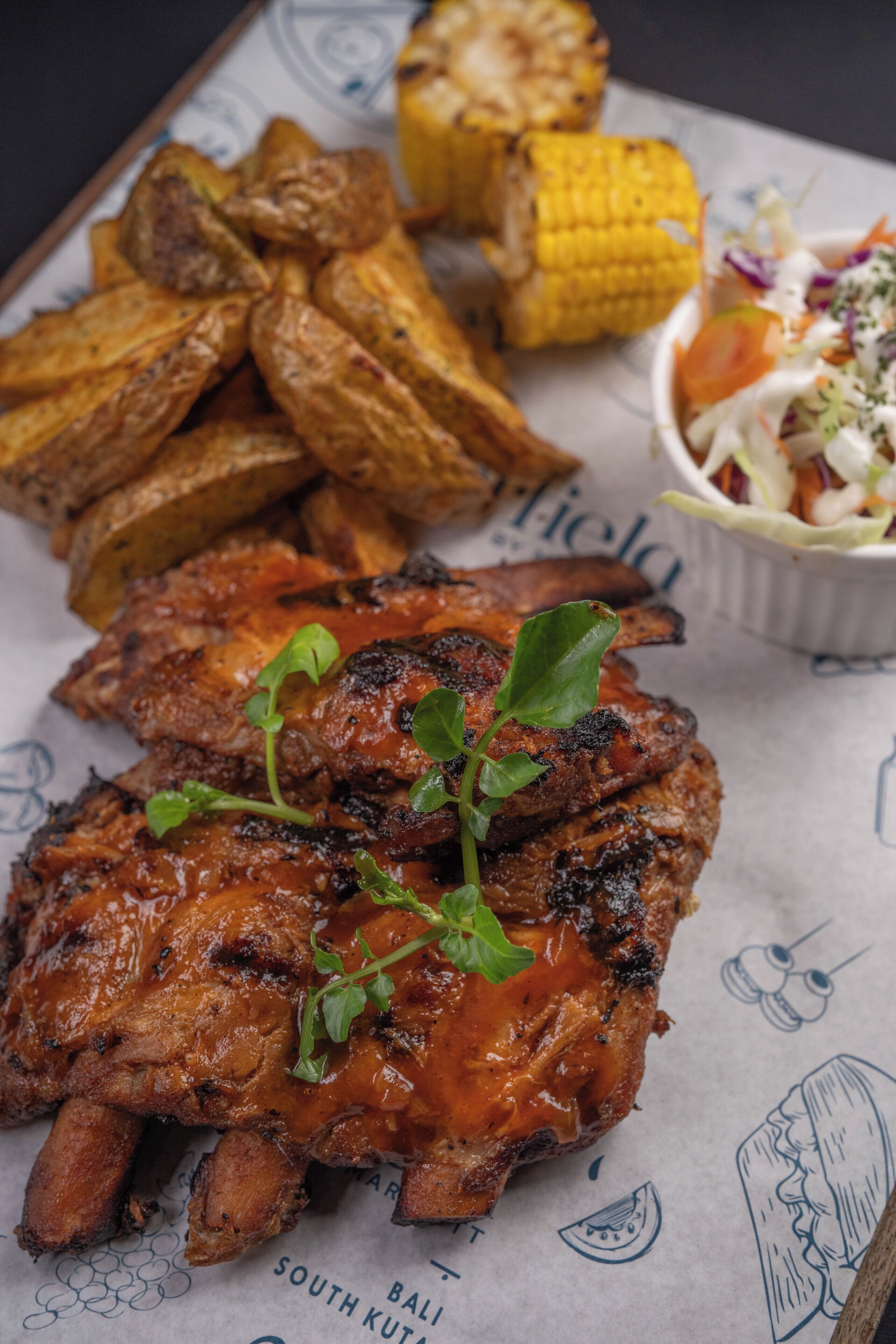 A plate of barbecued ribs with a side of roasted potatoes and a corn on the cob.