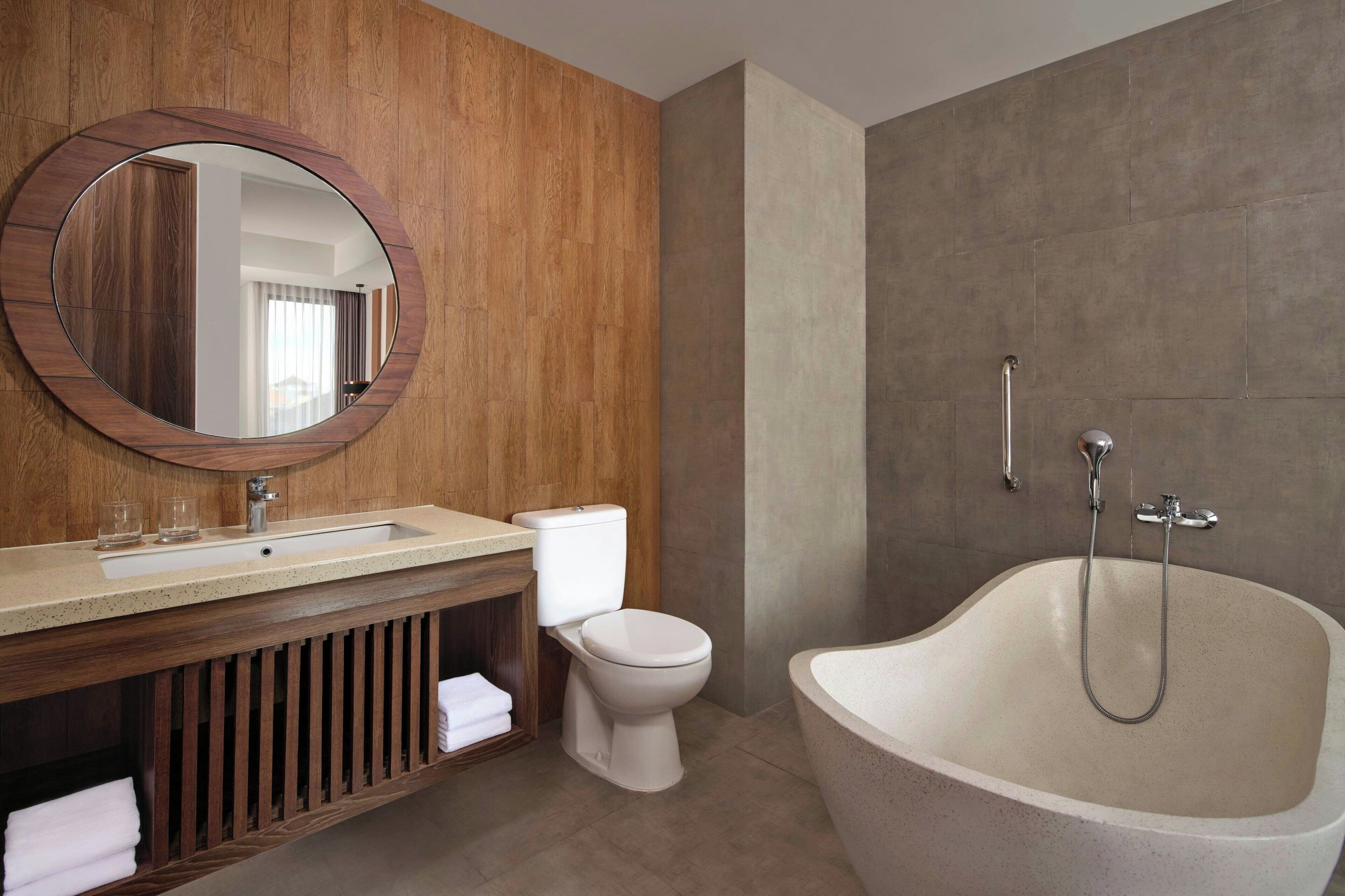 A modern bathroom with a wooden vanity, a round mirror, a toilet, and a freestanding bathtub.