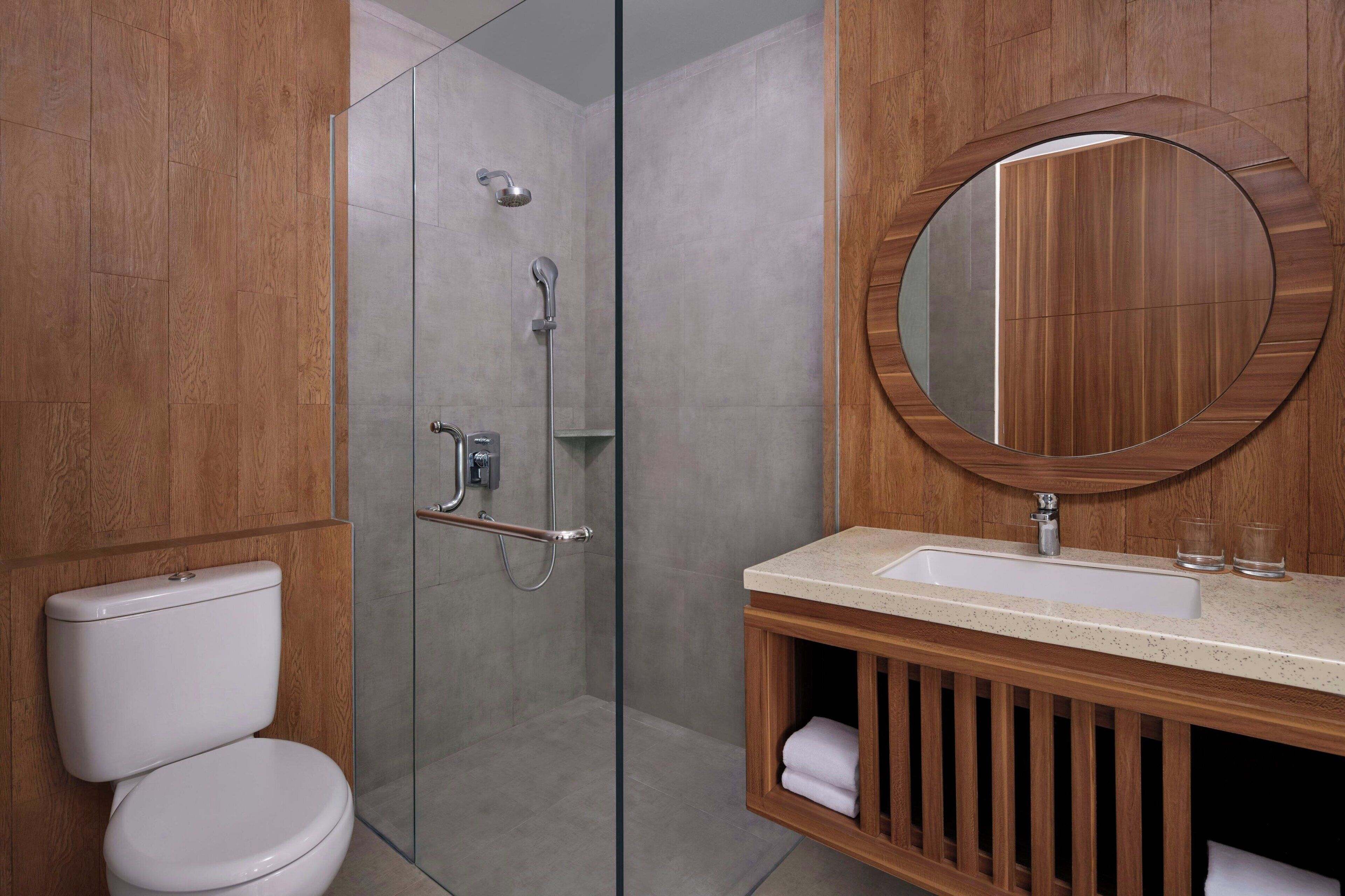 A modern bathroom with a glass shower enclosure, a wooden vanity with a round mirror, and a white toilet.