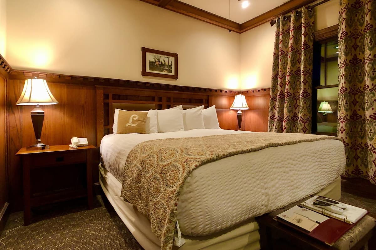 A hotel room with a large bed, two bedside lamps, a wooden headboard, a framed picture on the wall, and a window with curtains.
