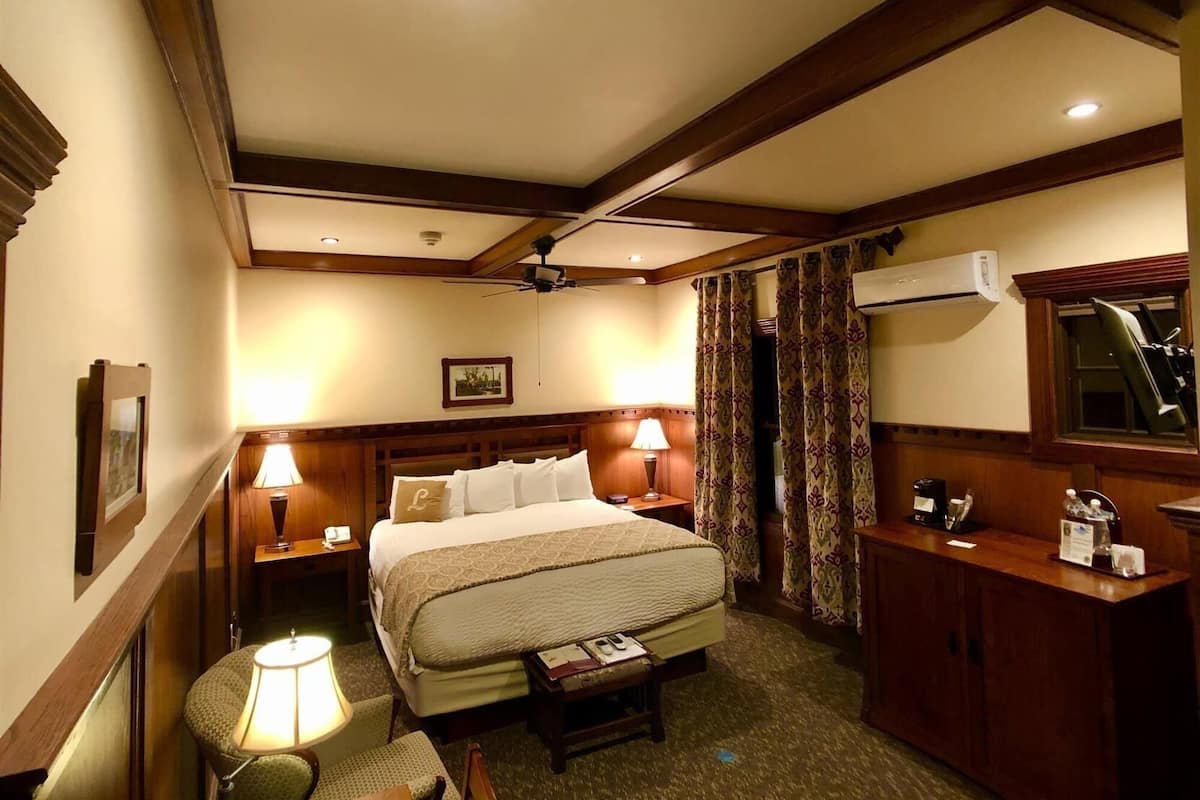 A hotel room with a large bed, two bedside lamps, a desk with a chair, an air conditioning unit, and a ceiling fan.