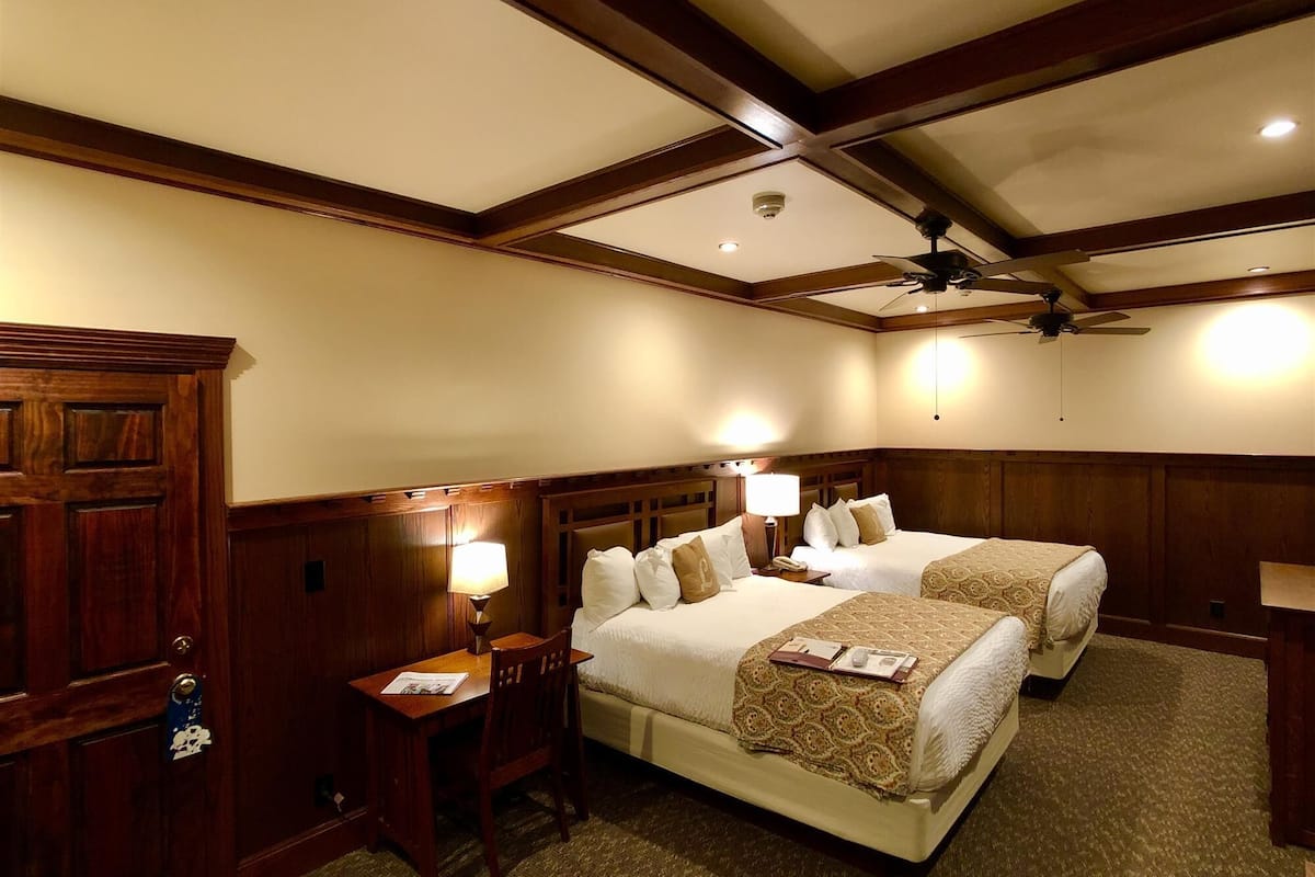 A hotel room with two beds, wooden paneling, a ceiling fan, and a desk with a lamp.