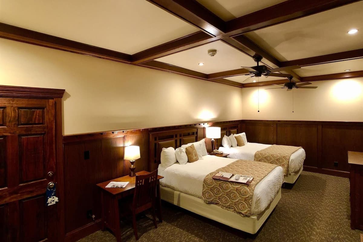 A hotel room with two beds, a desk, a chair, a ceiling fan, and a wooden door.