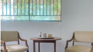 Two wooden chairs with beige cushions, a small round table with a white candle and a cup, and a window with green metal bars and a view of trees. - La Casa Verde x Townhouse in Mermoz, Dakar (Dakar)
