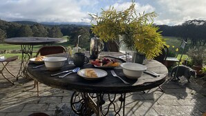 Ein runder Holztisch mit Blick auf eine grüne Landschaft, ein Metallkrug, ein Glas und ein Teller mit Essen.