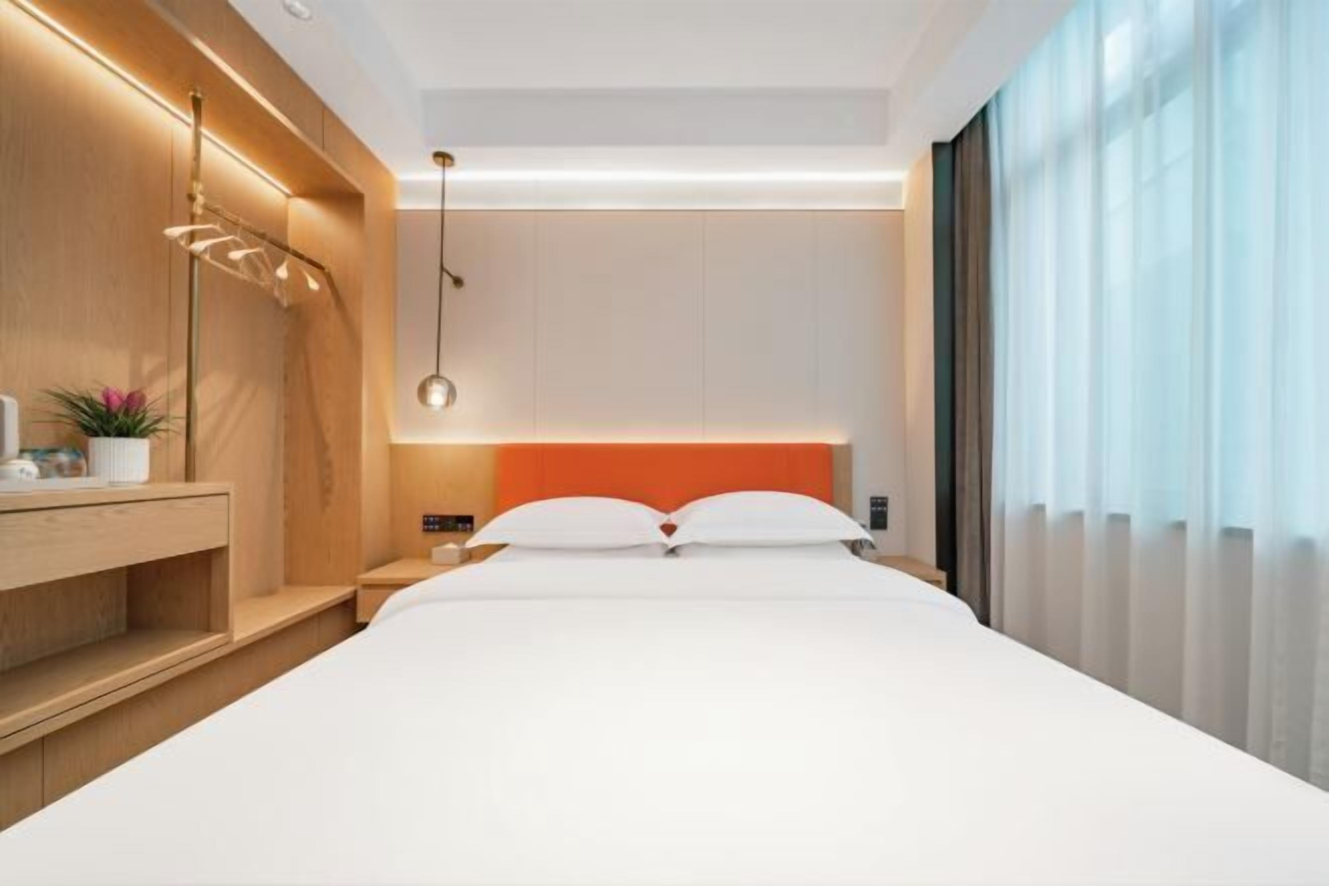 Une chambre d’hôtel moderne avec un grand lit, une table de chevet en bois, un luminaire fixé au mur et une grande fenêtre ornée de rideaux légers.