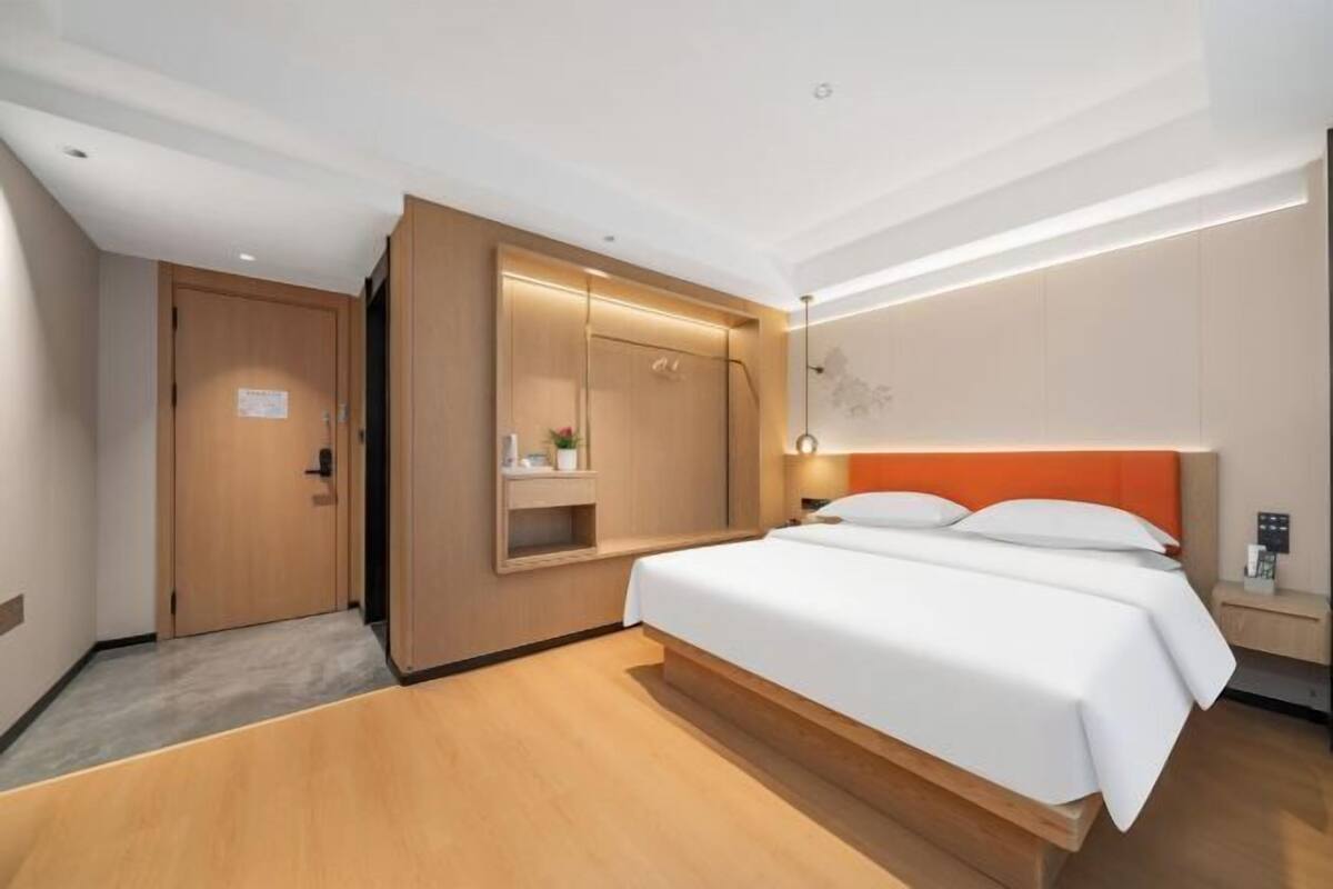 A modern hotel room with a wooden floor, a bed with white bedding, a wooden wardrobe, and a small bedside table with a lamp.