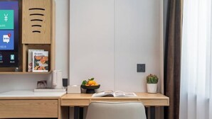 Una habitación de hotel moderna con un escritorio de madera, un monitor de ordenador, un libro, un frutero y una planta en maceta.