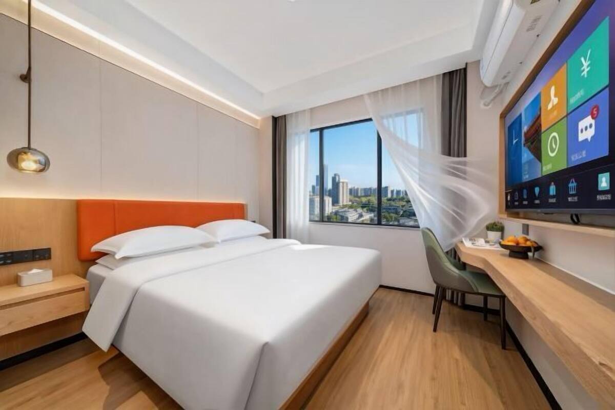 A modern hotel room with a bed, a desk, a chair, a TV, and a window with a city view.